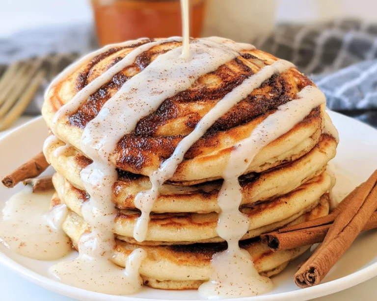 Stack of cinnamon roll pancakes drizzled with cream cheese glaze and served with cinnamon sticks.